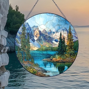 May include: A round stained glass window depicting a mountain lake scene. The artwork features a blue lake, green trees, and snow-capped mountains. The window is suspended by a chain and hangs in front of a scenic outdoor view.