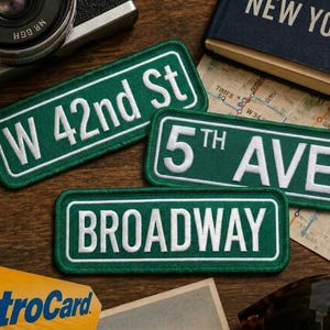 May include: Three rectangular embroidered patches featuring white text on a green background. The patches display the street names W 42nd St, 5th AVE, and BROADWAY in a style mimicking New York City street signs with white borders.