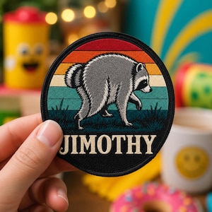 May include: A circular embroidered patch featuring a gray raccoon walking across a landscape with a retro sunset background of red, orange, and teal stripes. The word JIMOTHY is stitched in bold, cream-colored capital letters at the bottom.