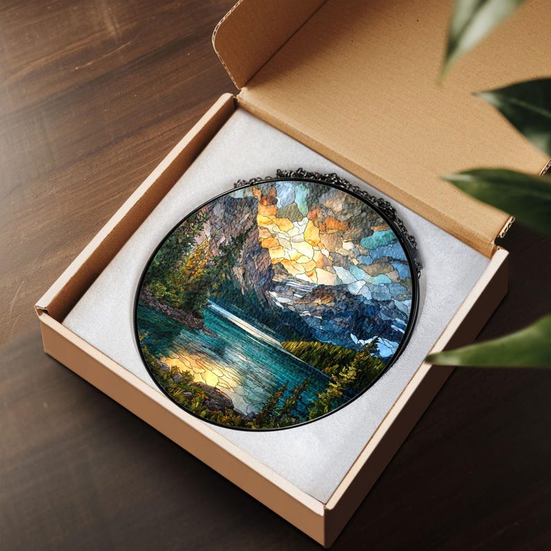 May include: A round stained glass art piece depicting a mountain lake scene, set in a black metal frame. The artwork features a vibrant color palette of blues, greens, oranges, and yellows, and is presented in a cardboard box with white padding.