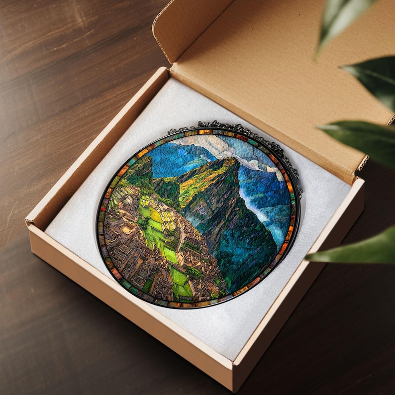 May include: A round stained glass art piece depicting a mountain landscape, presented in a cardboard box. The artwork features vibrant colors of green, blue, and brown, with a black metal frame. The box is open, revealing the art piece nestled in white padding.