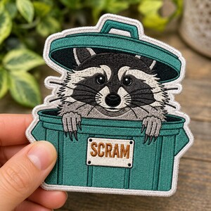 May include: An embroidered patch featuring a raccoon with black and white fur peeking out of a teal trash can with a lid. The raccoon has its paws resting on the rim of the bin. A white rectangular sign on the front of the bin reads SCRAM in brown.