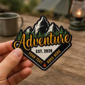 May include: A diamond-shaped embroidered patch featuring a mountain range with snow-capped peaks and green pine trees. The text reads Adventure in yellow script, with EST. 2026 and YOUR TEXT GOES HERE in black, plus a small orange flame icon.
