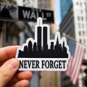 May include: A black and white embroidered patch featuring a silhouette of the New York City skyline, including the Twin Towers, with the text NEVER FORGET stitched in black capital letters on a white background at the bottom of the patch.