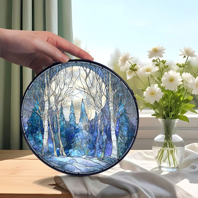 Op de afbeelding: Rond gebrandschilderd glaskunstwerk met een winterbossc&egrave;ne met volle maan. Het kunstwerk gebruikt blauw, wit en grijs, met gedetailleerde bomen en een besneeuwd landschap. Het kunstwerk wordt door een hand vastgehouden.