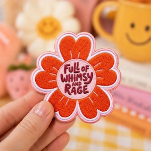 May include: A flower-shaped embroidered patch with bright orange petals, a light pink center, and a white border. The text in the center reads FULL OF WHIMSY AND RAGE in dark red lettering. The patch features a stitched texture throughout.
