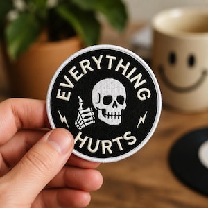 May include: A circular embroidered patch with a black background and a white border. The design features a white skull giving a thumbs-up gesture, flanked by two small lightning bolts. The text "EVERYTHING HURTS" is arched around the skull.