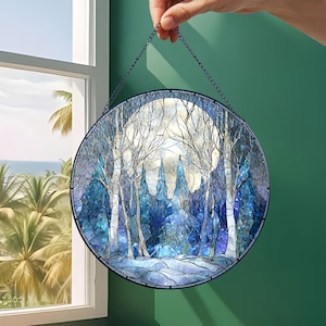 Op de afbeelding: Rond gebrandschilderd glaspaneel met een winterbossc&egrave;ne met volle maan. Het kunstwerk toont tinten blauw, wit en grijs, met bomen en een besneeuwd landschap. Het paneel wordt door een ketting vastgehouden.