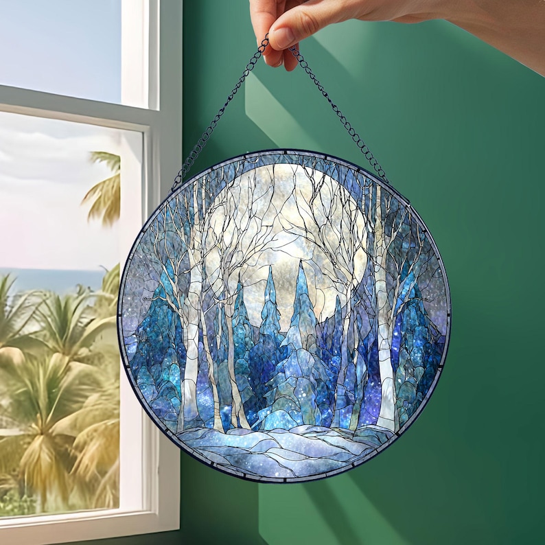 Op de afbeelding: Rond gebrandschilderd glaspaneel met een winterbossc&egrave;ne met volle maan. Het kunstwerk toont tinten blauw, wit en grijs, met bomen en een besneeuwd landschap. Het paneel wordt door een ketting vastgehouden.