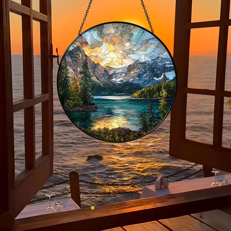 May include: A round stained glass window depicting a mountain lake scene with a sunset. The window is framed in black and hangs in front of an open window overlooking the ocean. The colors include blues, greens, oranges, and yellows.