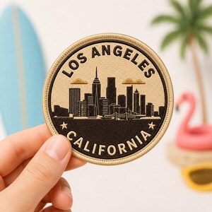 May include: A circular embroidered patch featuring a black skyline silhouette of Los Angeles, California. The design includes the text LOS ANGELES at the top and CALIFORNIA at the bottom, separated by two small stars, all on a tan background.