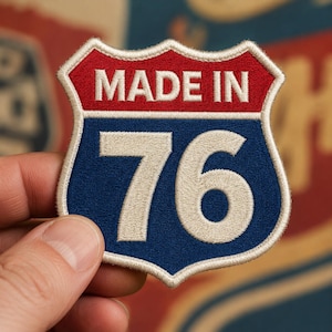 May include: A shield-shaped embroidered patch featuring a red top section with the white text MADE IN and a blue bottom section with the large white number 76. The entire patch is outlined with a thick cream-colored border and has a textured finish.