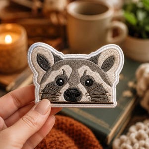 May include: An embroidered patch featuring the head of a raccoon with grey, white, and black thread detailing. The raccoon has large dark eyes, a black nose, and prominent whiskers, with a white border outlining the shape of the animal's head.