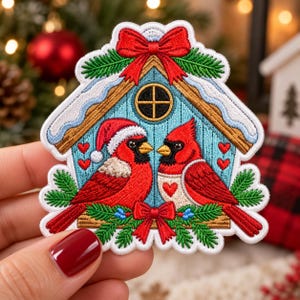 May include: An embroidered patch shaped like a house with a blue facade, featuring two red cardinal birds, one wearing a Santa hat. The design includes green pine branches, red bows, small red hearts, and a snow-covered roof with a round window.