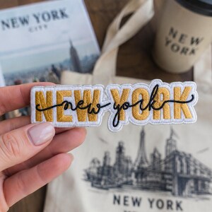 May include: An embroidered patch featuring the words NEW YORK in gold block letters with a white border, overlaid with the words new york in black cursive script. The patch is rectangular with a scalloped edge and a white fabric backing.