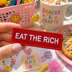 May include: A rectangular red embroidered patch with the white text EAT THE RICH stitched in a bold, sans-serif font. The patch features a clean, finished border and a dense, textured thread construction, presented as a flat, horizontal rectangular item.