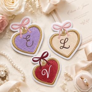 May include: Three heart-shaped embroidered patches featuring gold borders and a small bow at the top. The patches are purple with the letter E, cream with the letter L, and red with the letter V, each stitched in a cursive font style.