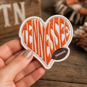 May include: A heart-shaped embroidered patch featuring the word TENNESSEE in white lettering against an orange background. The patch includes a small brown football graphic in the lower right corner and has a white stitched border edge.