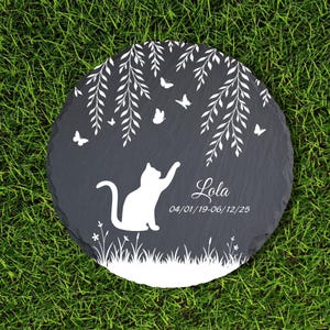 May include: A round, dark gray stone memorial with a white cat silhouette reaching for butterflies under a weeping willow design. The name "Lola" and dates are inscribed. The stone is set on green grass.