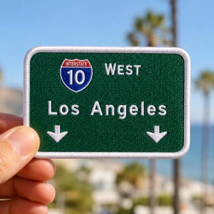 May include: A rectangular embroidered patch featuring a green background with a white border. It displays the Interstate 10 shield logo, the text WEST, the words Los Angeles, and two white downward-pointing arrows in a highway sign style.