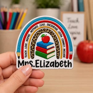May include: An embroidered patch featuring a rainbow arch with a leopard print band, a stack of three books, and a red apple. The text reads TEACH. LOVE. INSPIRE. MOTIVATE. LEAD. ENCOURAGE. LISTEN. CONNECT. INCLUDE and the name Mrs. Elizabeth.