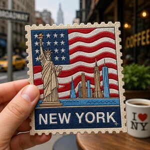 May include: A rectangular embroidered patch shaped like a postage stamp featuring the Statue of Liberty, a city skyline, and a wavy American flag background. The bottom blue border contains the white text NEW YORK with a small four-pointed star.