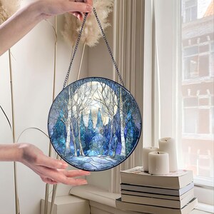 Op de afbeelding: Een rond gebrandschilderd glazen paneel met een winterbossc&egrave;ne met een volle maan. Het kunstwerk heeft tinten blauw, wit en grijs, met een zwarte metalen ketting om op te hangen. Het paneel wordt door een hand omhoog gehouden.