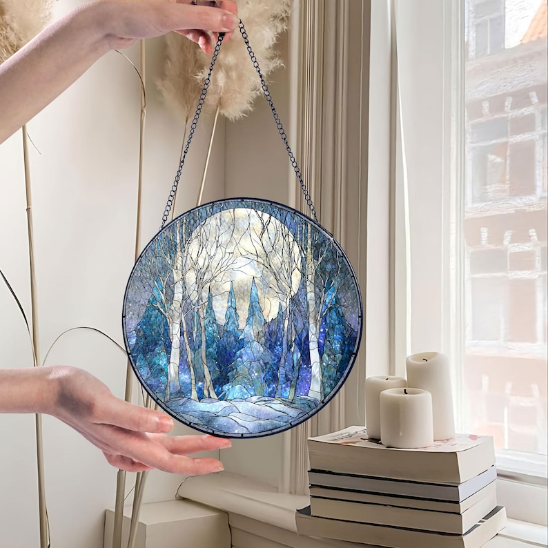 Op de afbeelding: Een rond gebrandschilderd glazen paneel met een winterbossc&egrave;ne met een volle maan. Het kunstwerk heeft tinten blauw, wit en grijs, met een zwarte metalen ketting om op te hangen. Het paneel wordt door een hand omhoog gehouden.