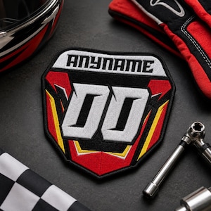 May include: A shield-shaped embroidered patch featuring the text ANYNAME in black on a white background, with large white numbers 00 centered below. The design includes red, black, and yellow geometric accents in a racing-inspired style.