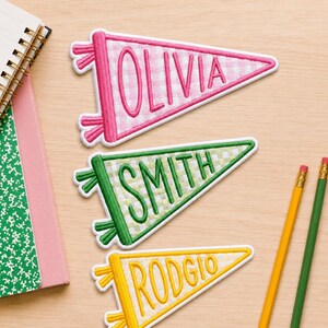May include: Three triangular pennant patches with embroidered borders and text. The top patch is pink with a white gingham pattern and the name OLIVIA. The middle is green with a white gingham pattern and SMITH. The bottom is yellow with RODGIO.