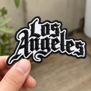 May include: An embroidered patch featuring the text Los Angeles in a bold, black gothic-style font with a white background and a thick black border. The lettering is stylized with sharp, decorative edges and a high-contrast color scheme.