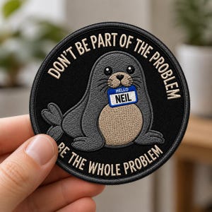 May include: A circular embroidered patch with a black background featuring a grey seal with a tan belly. The seal holds a blue and white name tag that reads HELLO NEIL. The text DON'T BE PART OF THE PROBLEM BE THE WHOLE PROBLEM is stitched in white.