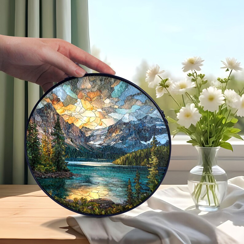 May include: A round, stained-glass art piece depicting a mountain lake scene with a vibrant sunset. The artwork features a black frame and is held by a hand. White flowers in a vase are in the background.