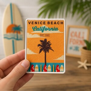 May include: A rectangular embroidered patch featuring a sunset scene with a palm tree, surfboards, and the text VENICE BEACH, California, and EST. 1905. The design uses orange, teal, white, and brown thread with a white stitched border.