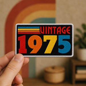 May include: A rectangular embroidered patch with a black background featuring the text VINTAGE in red and the number 1975 in large, multi-colored digits. The design includes horizontal stripes in blue, yellow, orange, and red on the left side.