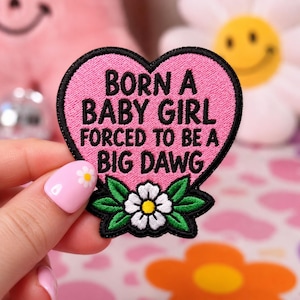May include: A heart-shaped embroidered patch with a pink background and a black border. The text reads BORN A BABY GIRL FORCED TO BE A BIG DAWG in black capital letters. At the bottom, there is a white daisy flower with green leaves.