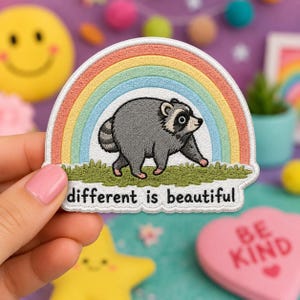 May include: An embroidered patch featuring a gray raccoon walking on green grass in front of a colorful rainbow. The text "different is beautiful" is stitched in black letters at the bottom of the patch, which has a white border.