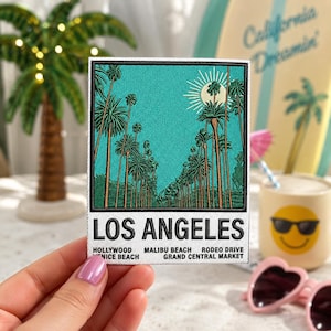May include: A rectangular embroidered patch featuring a teal sky with a sun and palm trees. Below the image, the text reads LOS ANGELES, HOLLYWOOD, MALIBU BEACH, RODEO DRIVE, VENICE BEACH, and GRAND CENTRAL MARKET in black capital letters on a white background.