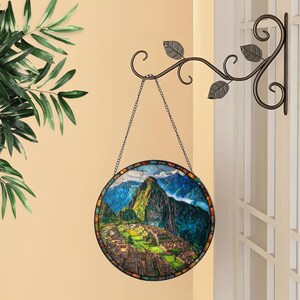 May include: A round stained glass panel depicting the Machu Picchu mountain landscape. The panel is framed with a mosaic border and hangs from a decorative metal chain. The panel is suspended from a decorative metal wall bracket with leaf accents.