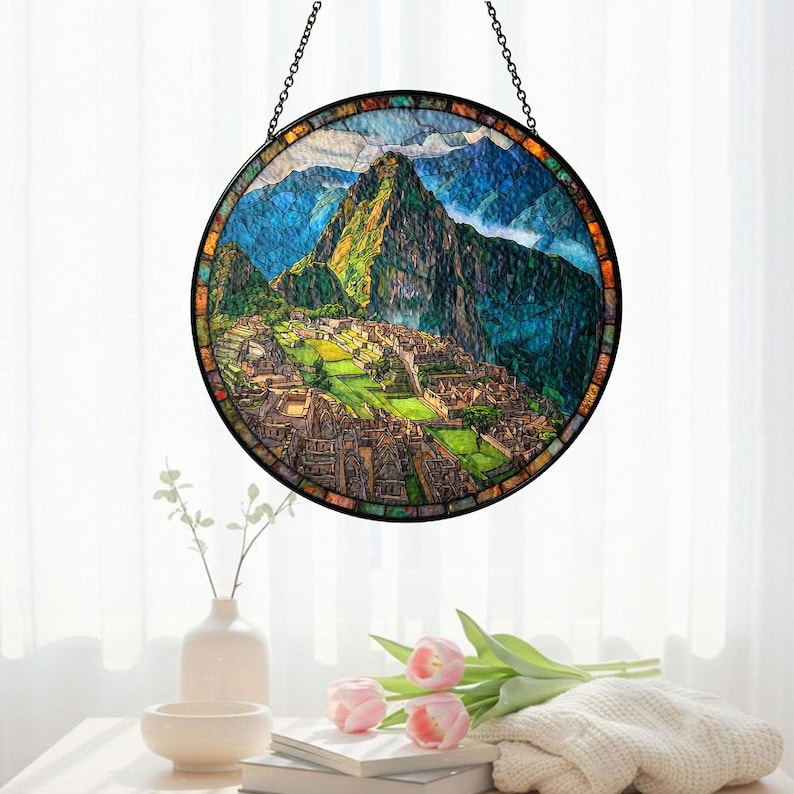 May include: Round stained glass window depicting Machu Picchu, with a black metal frame and hanging chain. The artwork features a colorful landscape of mountains, buildings, and sky. The window is displayed in front of a white curtain.