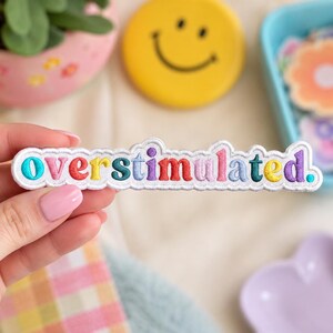 May include: A white embroidered patch with a scalloped edge featuring the word overstimulated. in colorful, multi-toned thread. Each letter is stitched in a different vibrant hue, including shades of blue, red, yellow, green, and purple.