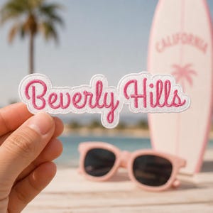 May include: An embroidered patch with a white background and pink cursive text that reads Beverly Hills. The patch has a die-cut shape that follows the contour of the lettering, featuring a clean white border and vibrant pink thread.