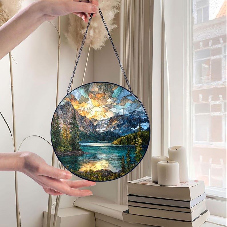 May include: Round stained glass panel depicting a mountain lake scene with a colorful sky. The artwork is framed in black and suspended by a chain. The panel features a vibrant landscape with trees, mountains, and water.