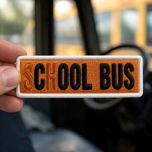 May include: A rectangular embroidered patch with a vibrant orange background and a thick white border. The text SCHOOL BUS is stitched in bold black capital letters across the center of the patch, featuring a clean and uniform design.