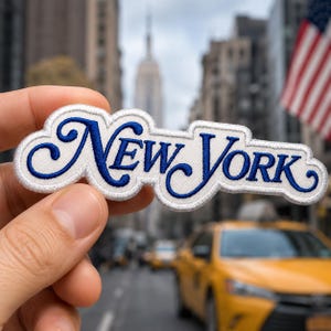 May include: An embroidered fabric patch featuring the text New York in a blue cursive script font with a white background and a white border. The patch has a die-cut shape that follows the contours of the lettering, creating a distinct outline.