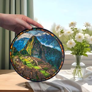 May include: Round stained glass art piece depicting Machu Picchu, with a black frame. The artwork features a colorful mosaic of the mountain landscape, with green, blue, and brown hues. The piece is held in a hand, with a vase of white flowers in the background.