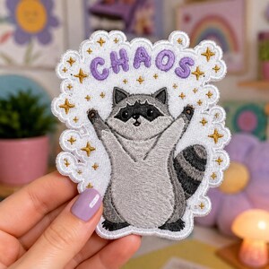 May include: An embroidered patch featuring a gray and white raccoon with its arms raised, surrounded by gold embroidered stars on a white background. The word CHAOS is stitched in purple letters at the top. The patch has a scalloped white border.