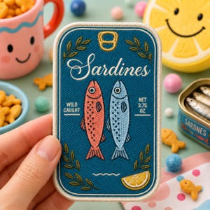 May include: A rectangular embroidered patch featuring a dark blue background with the text Sardines, Wild Caught, and Net 3.75 oz. The design includes a coral and a light blue fish, a yellow lemon slice, green leaf accents, and a gold pull tab.