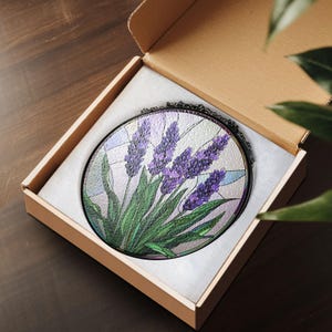 May include: A round stained glass suncatcher with a lavender flower design. The suncatcher is in a brown cardboard box with white tissue paper.