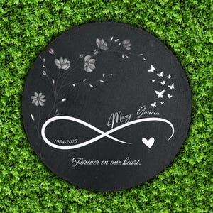 May include: A round, black memorial stone with white floral and butterfly designs. The name "Mary Garcia" is inscribed, along with the years "1984-2025" and the phrase "Forever in our heart."